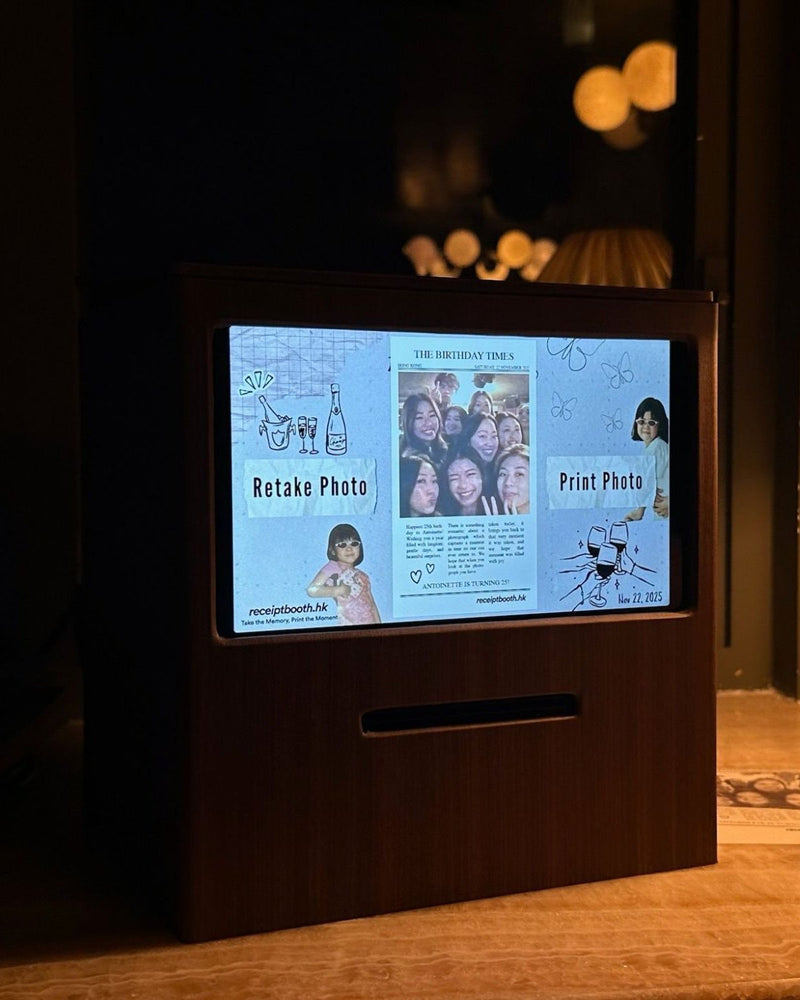 Walnut-finish Receipt Booth in a dimly lit room, its screen showing a custom newspaper-style template around a group photo, with a printed strip on the table beside it