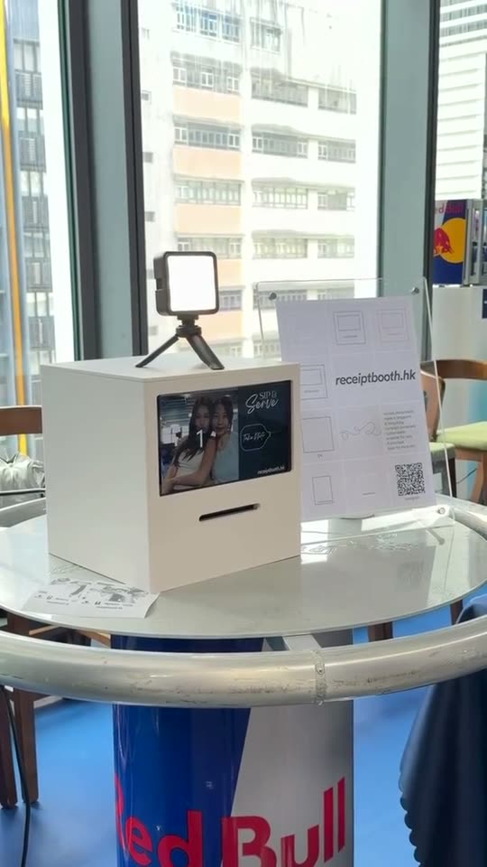 Cream Receipt Booth with a small light on top, on a high round table by a window, as guests tap its screen and printed strips lie on the table